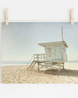 A photo of Offley Green's California Summer Beach Art, 'Malibu Lifeguard Tower', numbered "3" on Malibu beach, pinned by wooden clothespins on a line, set against a scene with soft sand and a clear sky.