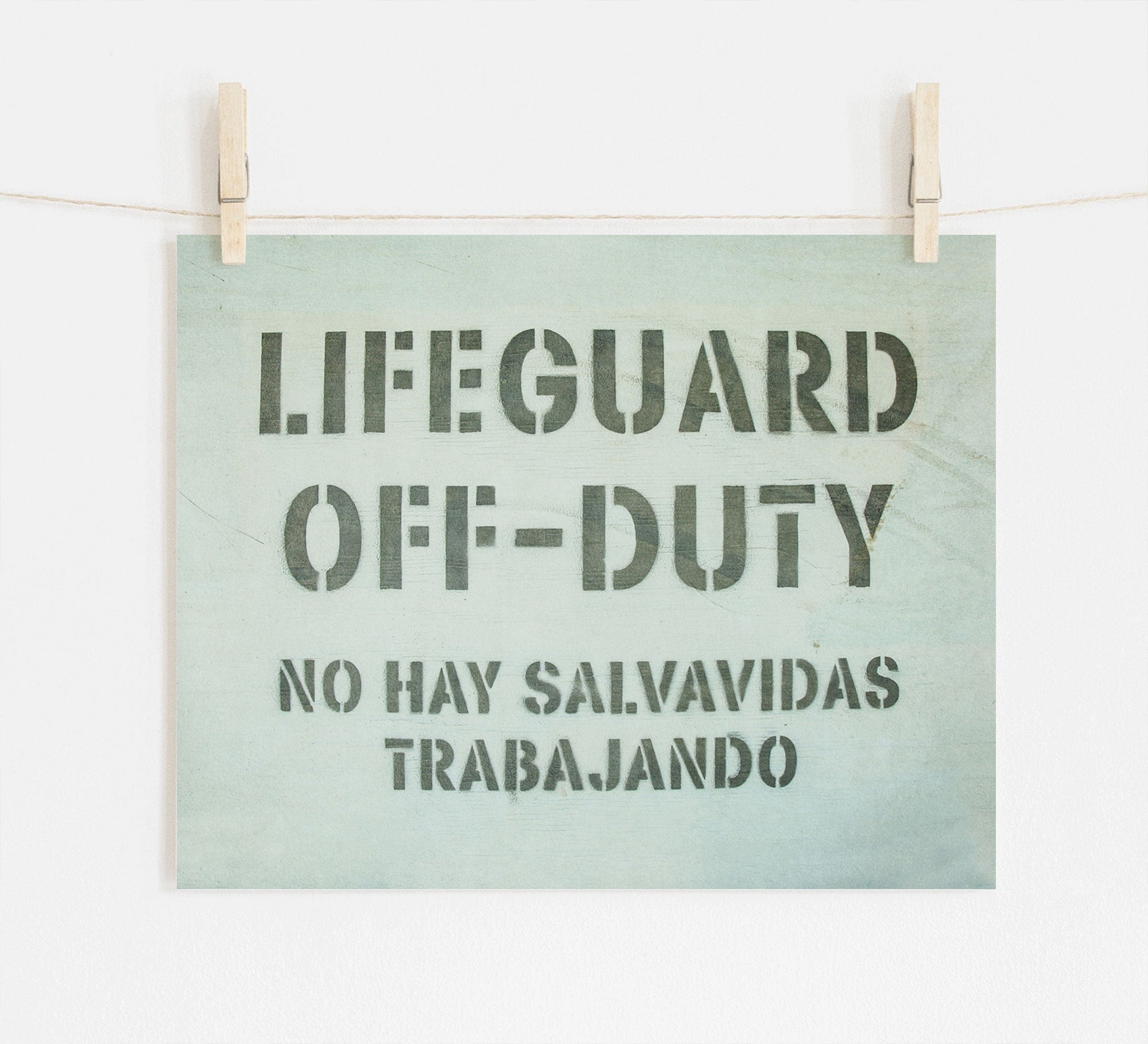 A sign hangs from two clothespins on a string, reading "LIFEGUARD OFF-DUTY" in large letters with "NO HAY SALVAVIDAS TRABAJANDO" below. The playful wall art, reminiscent of something you'd find near lifeguard towers, adds a charming touch to any space. This is the Teal Blue Surfer Beach Print, 'Off Duty' by Offley Green.