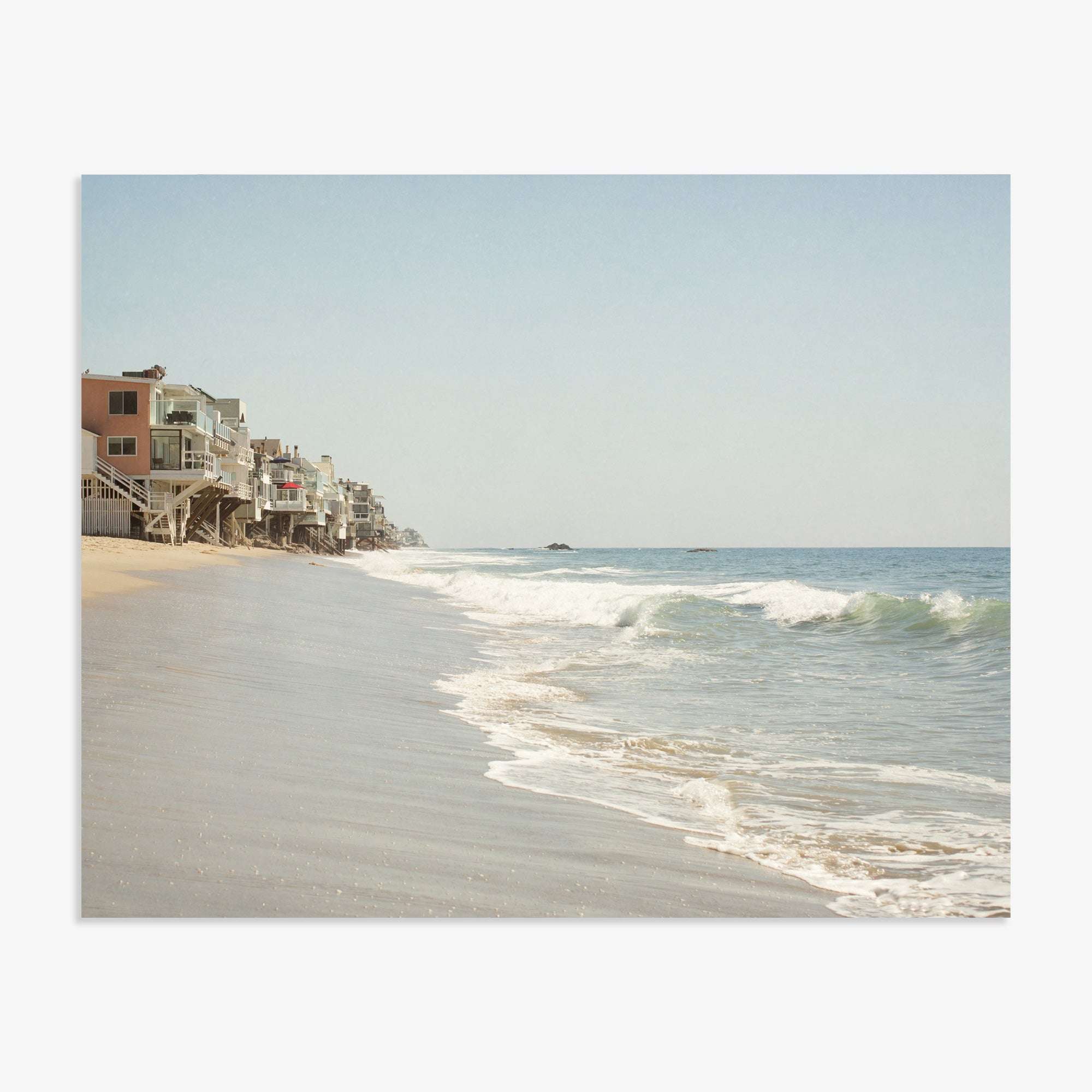A serene Malibu coastline scene with gentle waves lapping at the shore and a row of multistory beach houses on stilts, lining the sandy coast under a clear sky featuring the Malibu Beach House Print by Offley Green.
