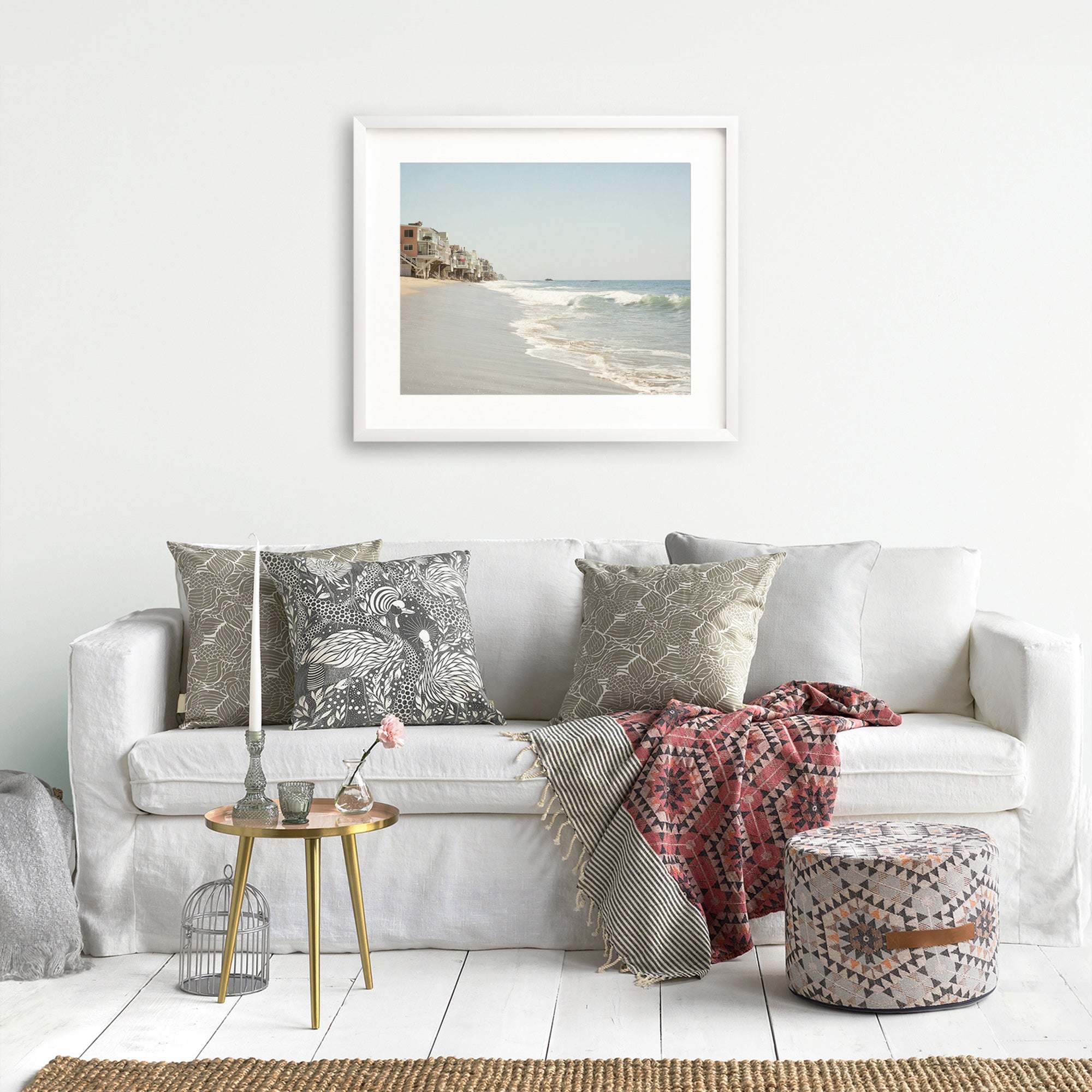 A cozy living room setup with a white sofa adorned with various patterned cushions, a throw blanket, a small round table with flowers, a cage-like decor item, and Offley Green's Malibu Beach House Print, 'Ocean View' wall art.