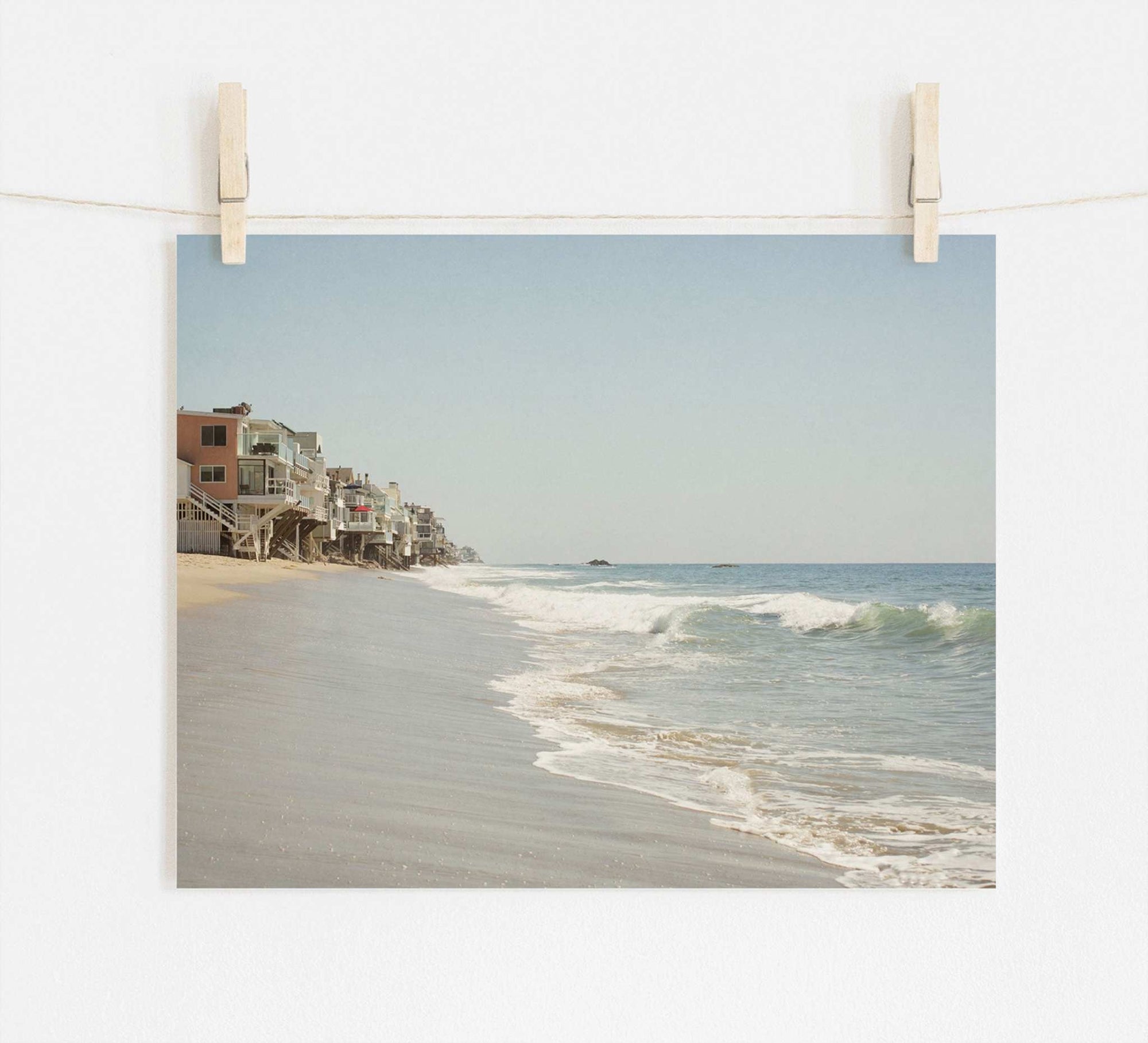 A photograph clipped to a string showing the serene Malibu coastline with waves gently lapping the shore and a row of Offley Green Malibu Beach House Print, 'Ocean View' built along a sandy beachfront.