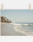 A photograph clipped to a string showing the serene Malibu coastline with waves gently lapping the shore and a row of Offley Green Malibu Beach House Print, 'Ocean View' built along a sandy beachfront.