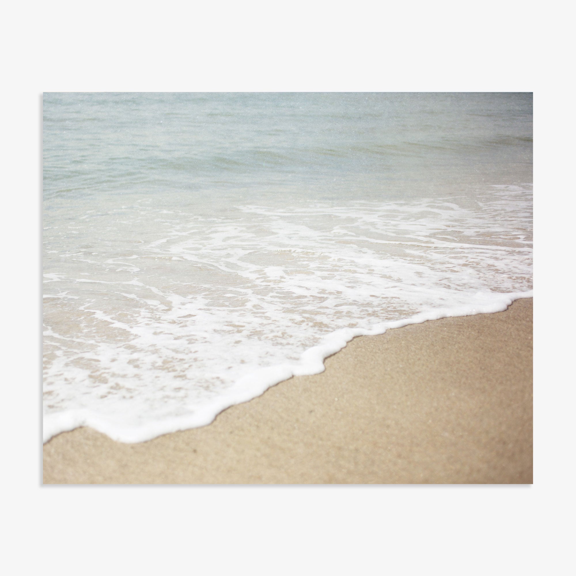 Gentle waves wash over a sandy beach in California, creating a soft, foamy edge where the water meets the shore, with a glimpse of the calm sea extending into the distance. Offley Green&#39;s Beach Waves Print, &#39;Chasing Surf&#39;.