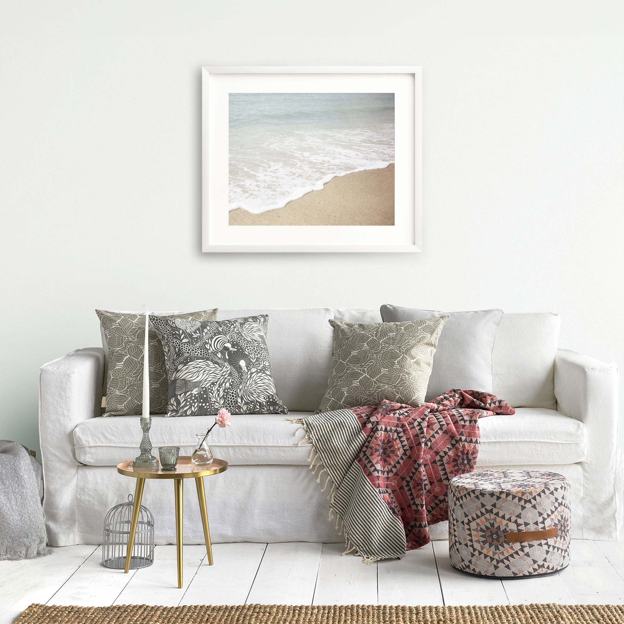 A cozy living room setup with a white sofa adorned with patterned cushions, a red throw blanket, a small round table with books, and an 'Offley Green' Beach Waves Print, 'Chasing Surf' hanging above.