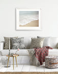 A cozy living room setup with a white sofa adorned with patterned cushions, a red throw blanket, a small round table with books, and an 'Offley Green' Beach Waves Print, 'Chasing Surf' hanging above.