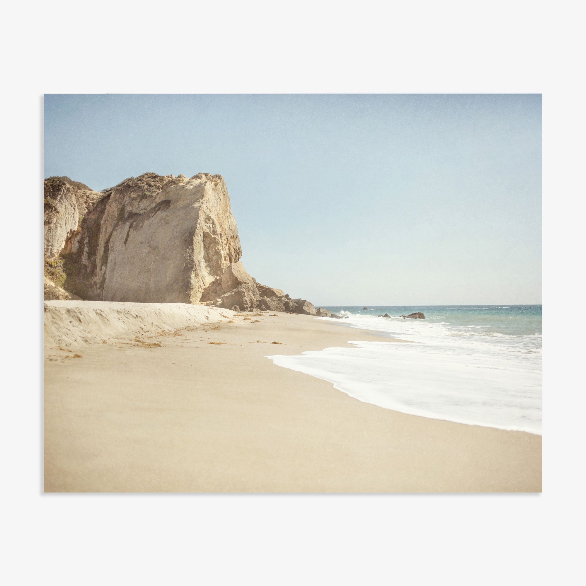 A serene Offley Green California Malibu Print, &#39;Point Dume&#39; beach scene with soft waves lapping at the shore, a large cliff to the left, and a clear blue sky. The setting conveys a peaceful, natural coastal landscape.