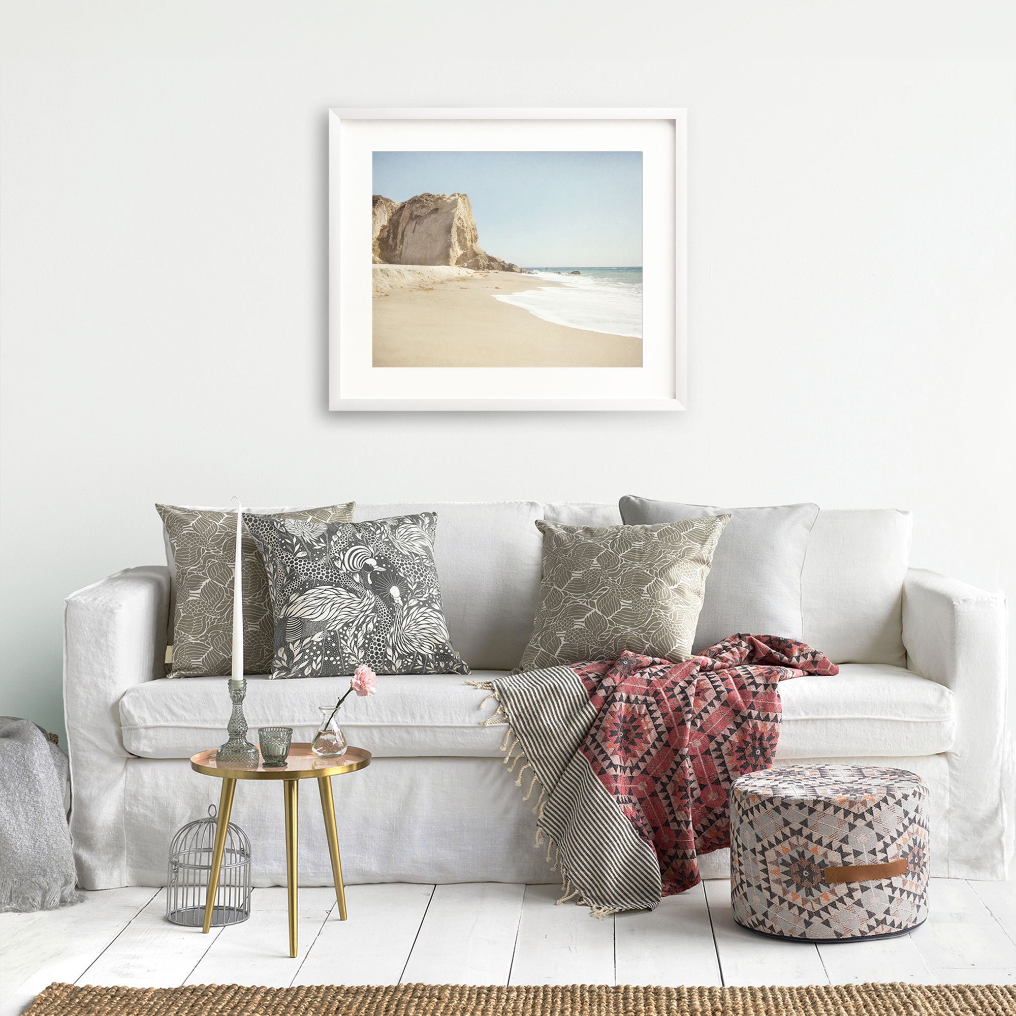 A cozy living room with a white couch adorned with decorative pillows, a framed Offley Green California Malibu Print, 'Point Dume' landscape on the wall, a small table with a vase next to the sofa, a blanket, and a pattern.