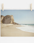 A photo of Offley Green's California Malibu Print, 'Point Dume' with waves gently lapping the shore and a large rocky outcrop to the left. The picture is pinned to a wall by wooden clothespins.