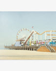 A seaside amusement park featuring a ferris wheel and roller coaster on the Santa Monica Pier, set against a clear sky. The sandy beach in the foreground is empty and expansive. Offley Green's California Print, 'Santa Monica Pier'.