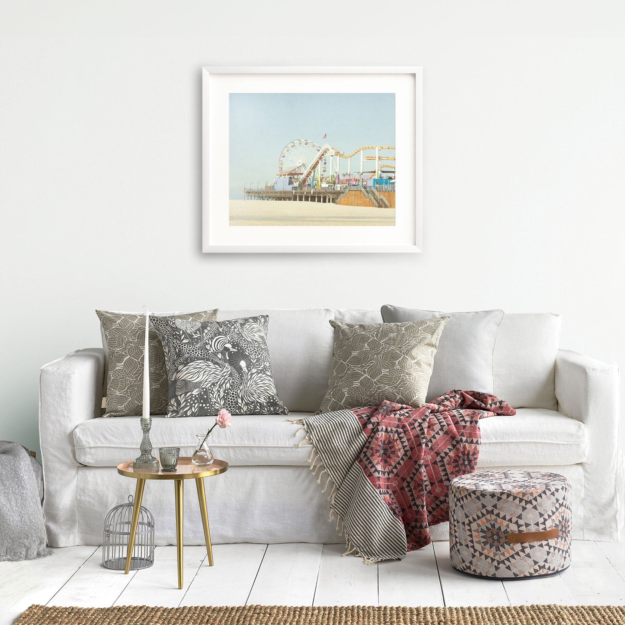 A cozy living room with a white sofa adorned with patterned pillows, a red throw blanket, a small round table, and a birdcage decor item, featuring a framed painting of the Offley Green California Print, 'Santa Monica Pier'