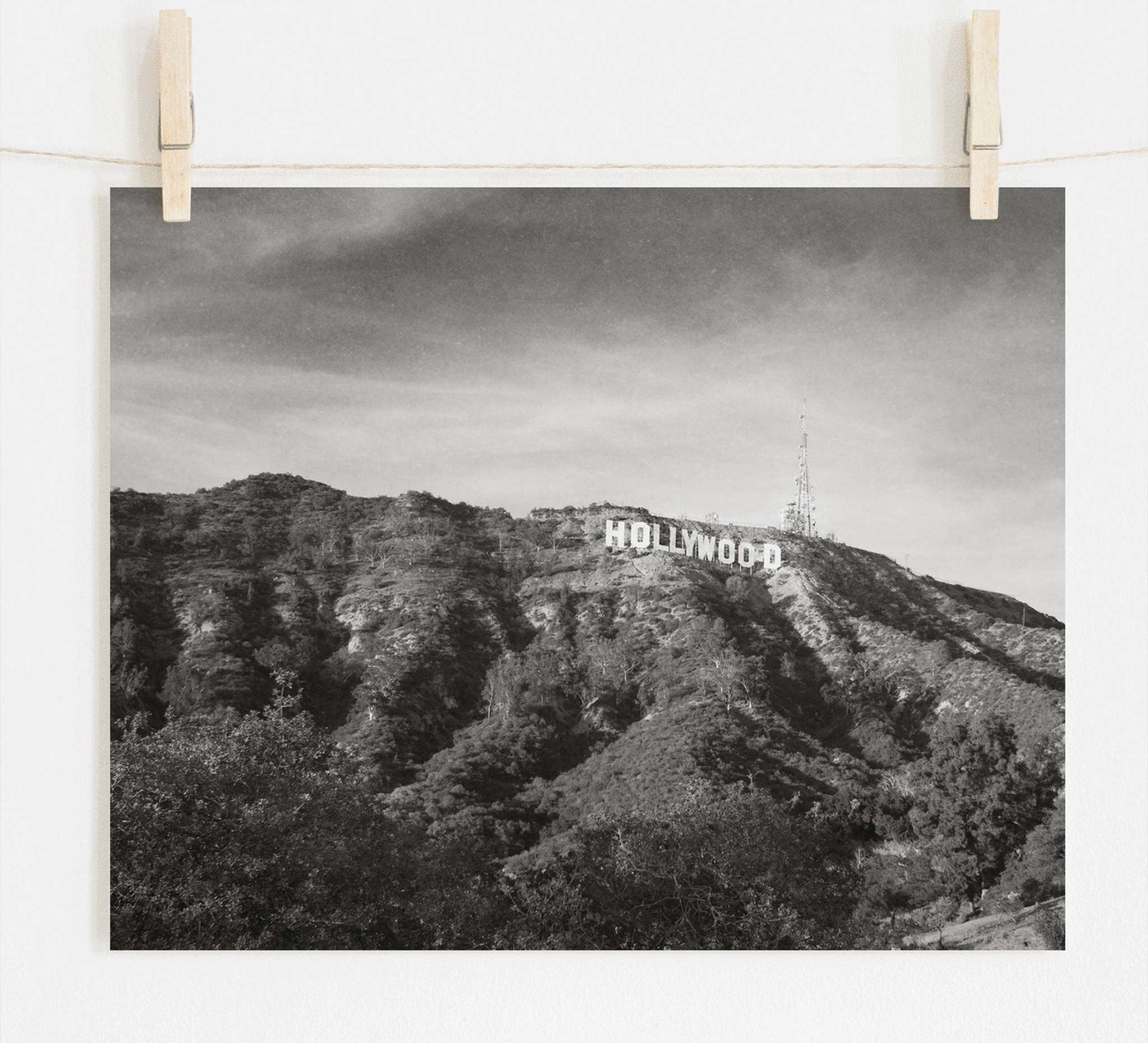 Black and white film noir photograph of the Offley Green Hollywood Sign Black and White Vintage Print, 'Old Hollywood' pinned up, set against a backdrop of a leafy hill with broadcasting antennas. The image has a rustic and vintage feel.