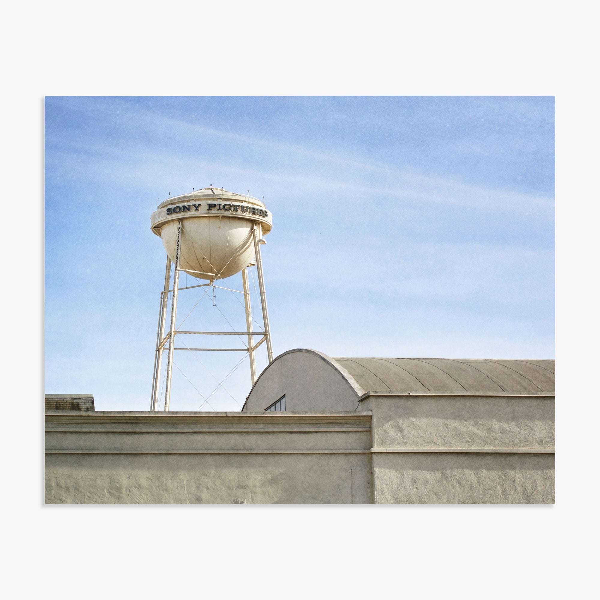 A white water tower labeled &quot;Offley Green Studios&quot; stands against a clear blue sky, partly obscured by the top of a building with a curved roof in the foreground.