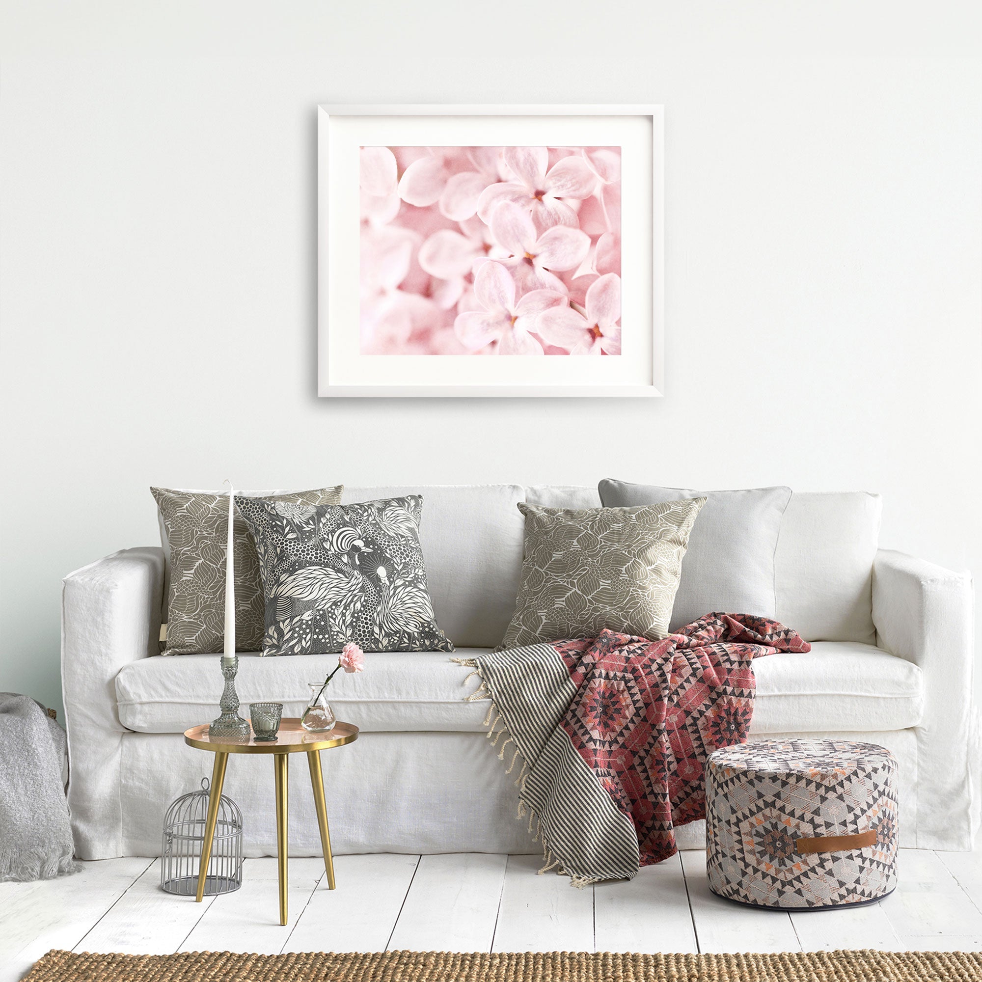 A cozy living room setup with a white sofa adorned with various patterned cushions, a knit throw blanket, and a small golden side table with decorative items, under Offley Green's Pink Botanical Print, 'Bed of Lilacs'.