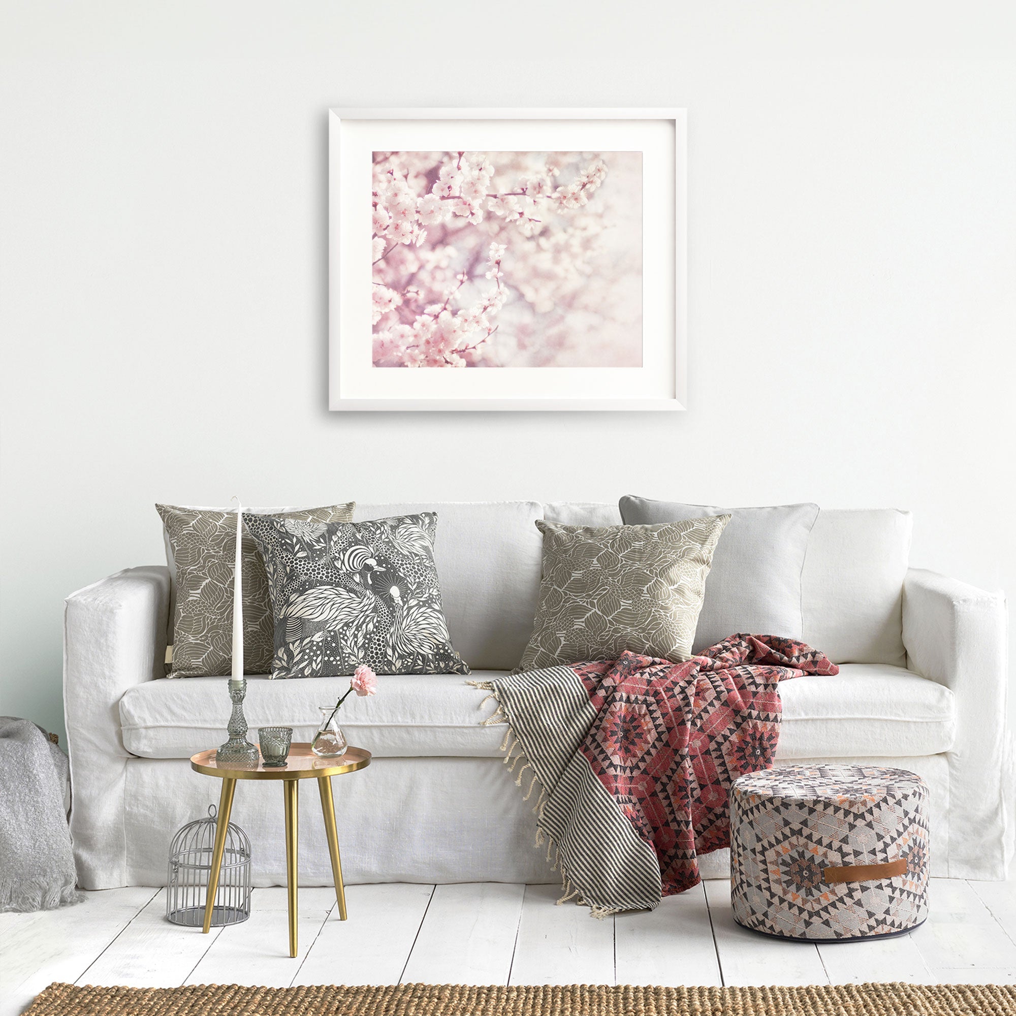 A cozy living room corner with a white sofa adorned with patterned cushions, a pink and gray throw blanket, a small round glass table, a decorative birdcage, and the Offley Green Pink Floral Print, &#39;Dreamy Blossom.&#39;