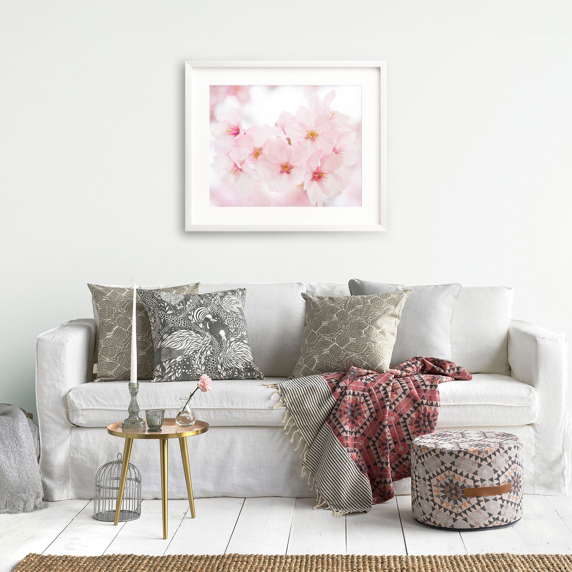 A cozy living room with a white sofa adorned with patterned cushions, a framed Pink Flower Print, 'Cherry Blossom' above it on archival photographic paper, a small round table with books, a birdcage, and a kn