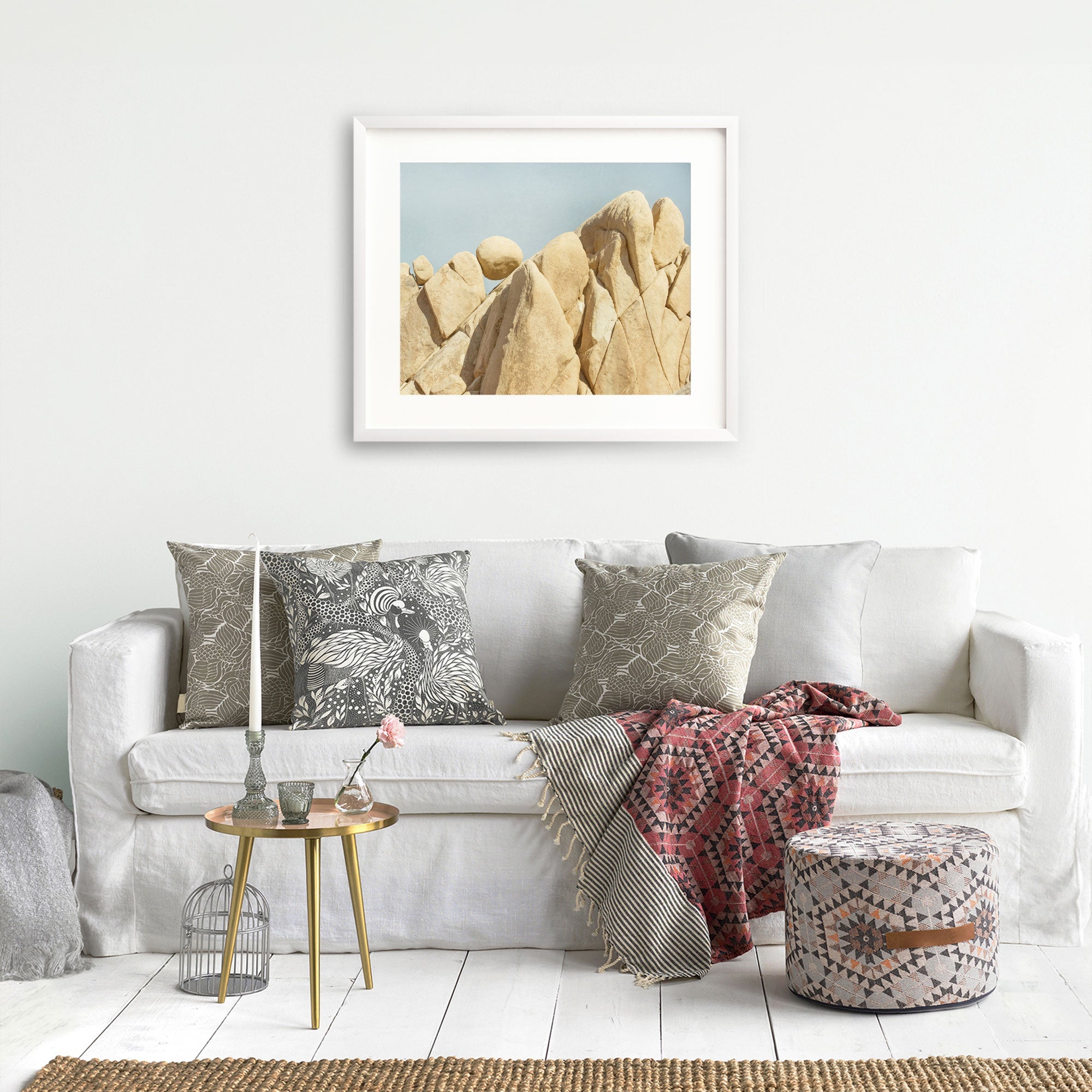 A cozy living room with a white sofa adorned with patterned cushions, a red patterned throw, a small coffee table with books and a vase, an unframed Offley Green print of Joshua Tree National Park &#39;Rock Formations&#39;.