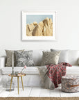 A cozy living room with a white sofa adorned with patterned cushions, a red patterned throw, a small coffee table with books and a vase, an unframed Offley Green print of Joshua Tree National Park 'Rock Formations'.