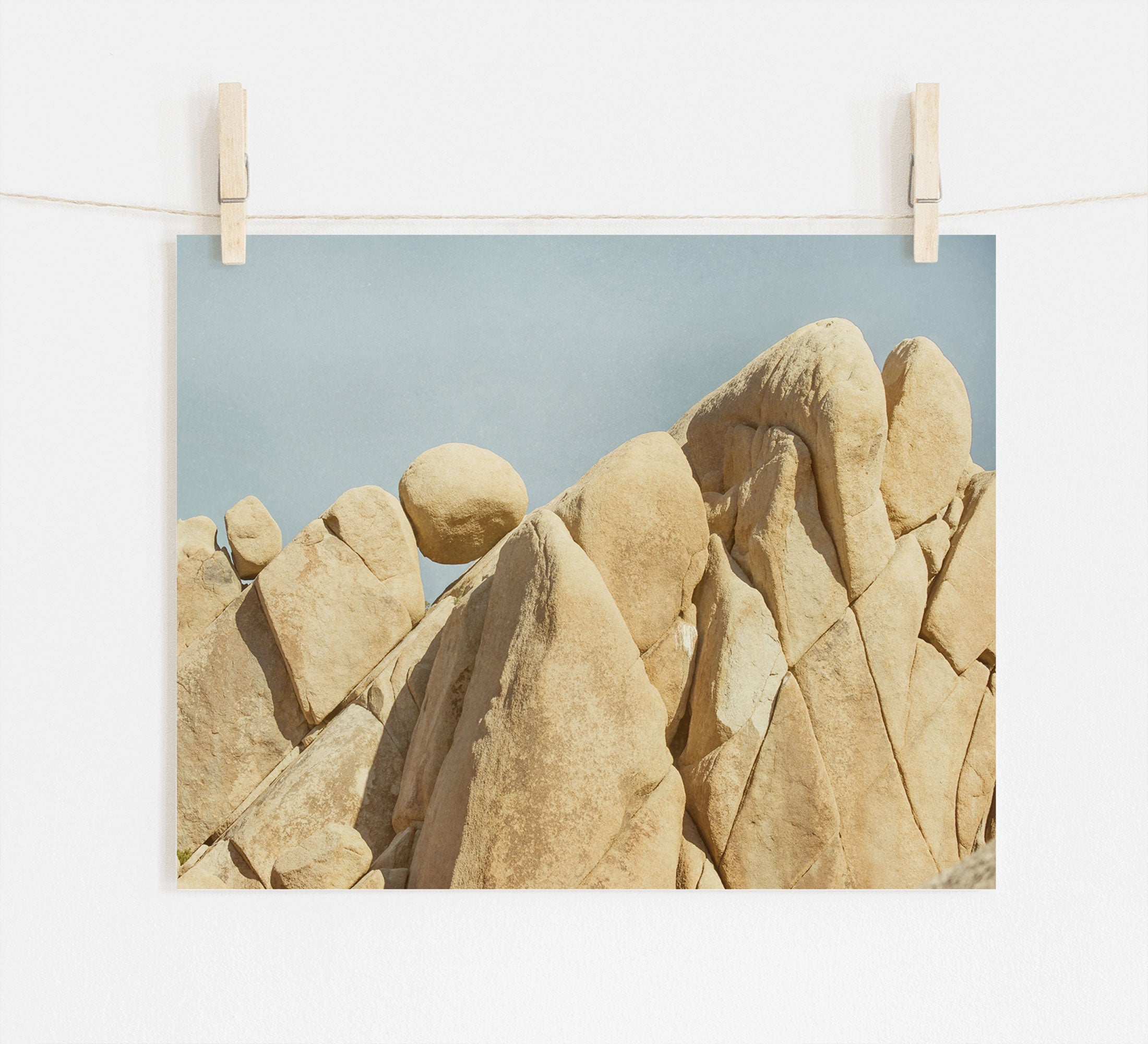 A photograph of rugged, beige rocks in Joshua Tree National Park against a clear blue sky, clipped to a string with two wooden clothespins, displayed against a white wall. Offley Green&#39;s Joshua Tree Print, &#39;Rock Formations.&#39;