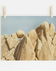 A photograph of rugged, beige rocks in Joshua Tree National Park against a clear blue sky, clipped to a string with two wooden clothespins, displayed against a white wall. Offley Green's Joshua Tree Print, 'Rock Formations.'