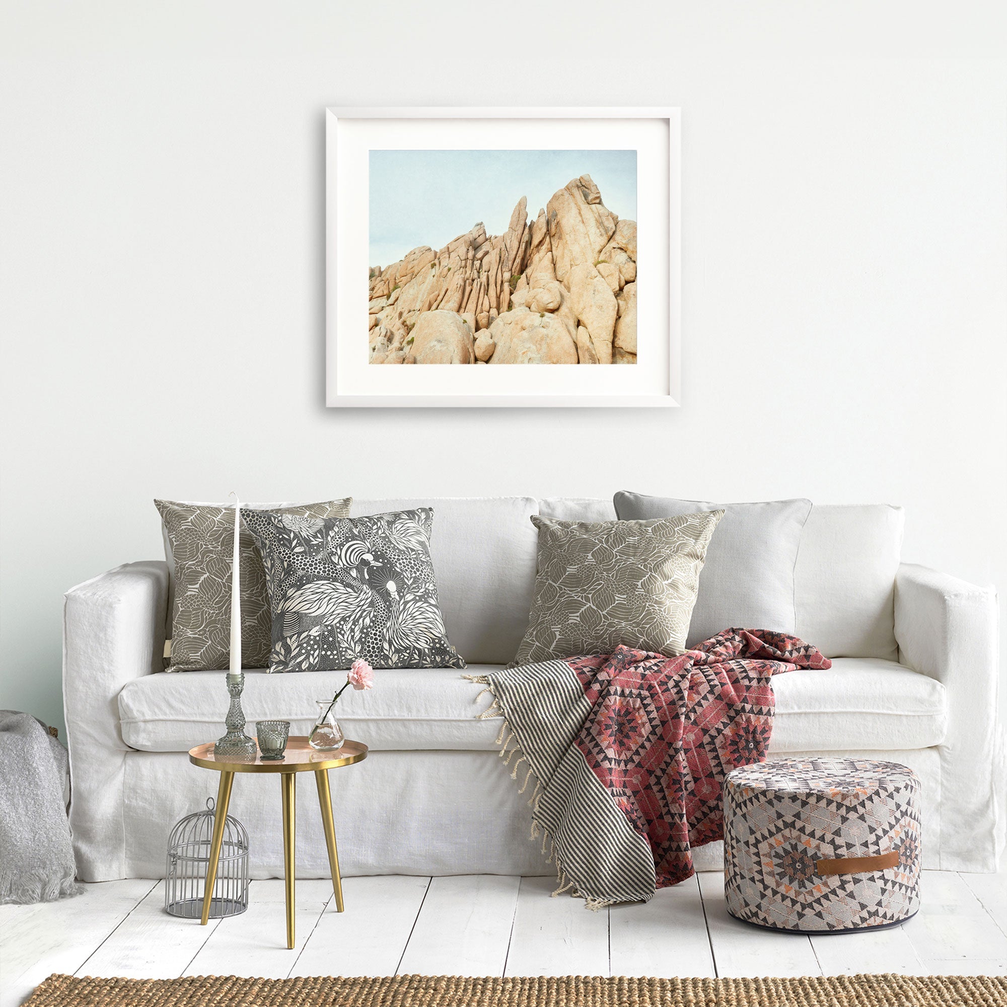 A cozy living room featuring a white sofa adorned with decorative gray pillows, a red patterned throw blanket, a small round table with a plant, a birdcage, and unframed prints of Offley Green's Joshua Tree Print, 'Joshua Rocks'.