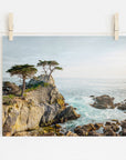 A serene photograph of California Coastal Print, 'Lone Cypress' from Offley Green with two trees atop a cliff overlooking the ocean, pinned to a white wall with wooden clothespins.
