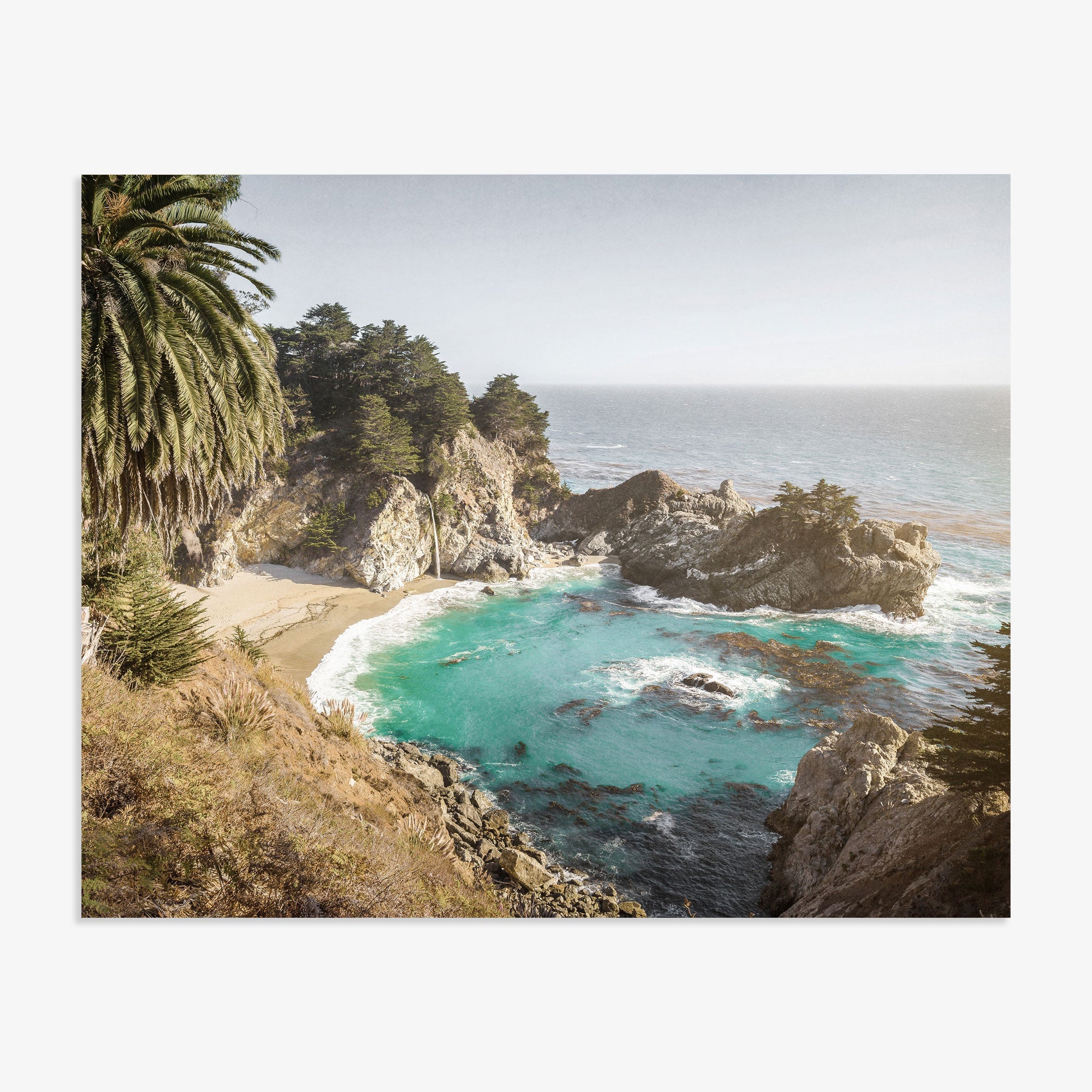 A scenic coastline with turquoise waters surrounded by rugged cliffs and lush greenery, featuring a prominent cove and a tall palm tree on the left, captures the essence of Offley Green&#39;s Big Sur Coastal Print, &#39;Julia Pffeifer&#39; wall art.