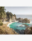 A scenic coastline with turquoise waters surrounded by rugged cliffs and lush greenery, featuring a prominent cove and a tall palm tree on the left, captures the essence of Offley Green's Big Sur Coastal Print, 'Julia Pffeifer' wall art.