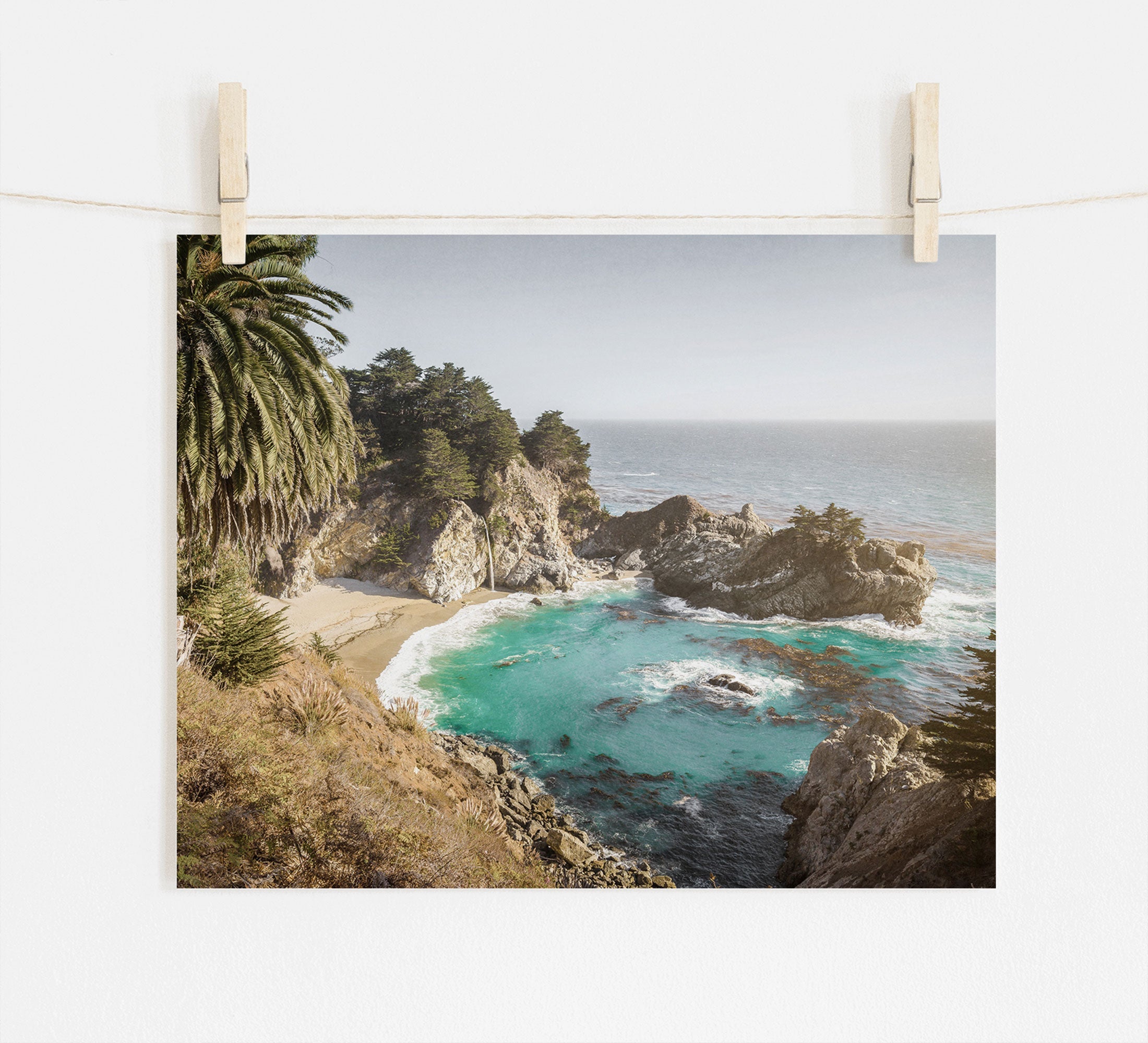 Photo of a scenic Offley Green Big Sur Coastal Print, 'Julia Pffeifer' with turquoise waters, suspended by clothespins. The view includes rocky cliffs, a sandy beach, and lush greenery under a bright sky.