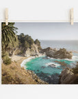 Photo of a scenic Offley Green Big Sur Coastal Print, 'Julia Pffeifer' with turquoise waters, suspended by clothespins. The view includes rocky cliffs, a sandy beach, and lush greenery under a bright sky.