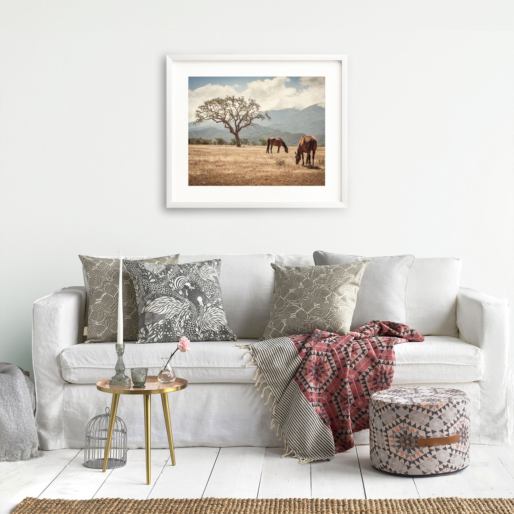 A cozy living room featuring a white sofa adorned with patterned cushions, a red throw, a small round table with decor items, and a framed rustic print of horses in a field titled 'Santa Ynez Horses' by Offley Green.