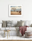A cozy living room featuring a white sofa adorned with patterned cushions, a red throw, a small round table with decor items, and a framed rustic print of horses in a field titled 'Santa Ynez Horses' by Offley Green.