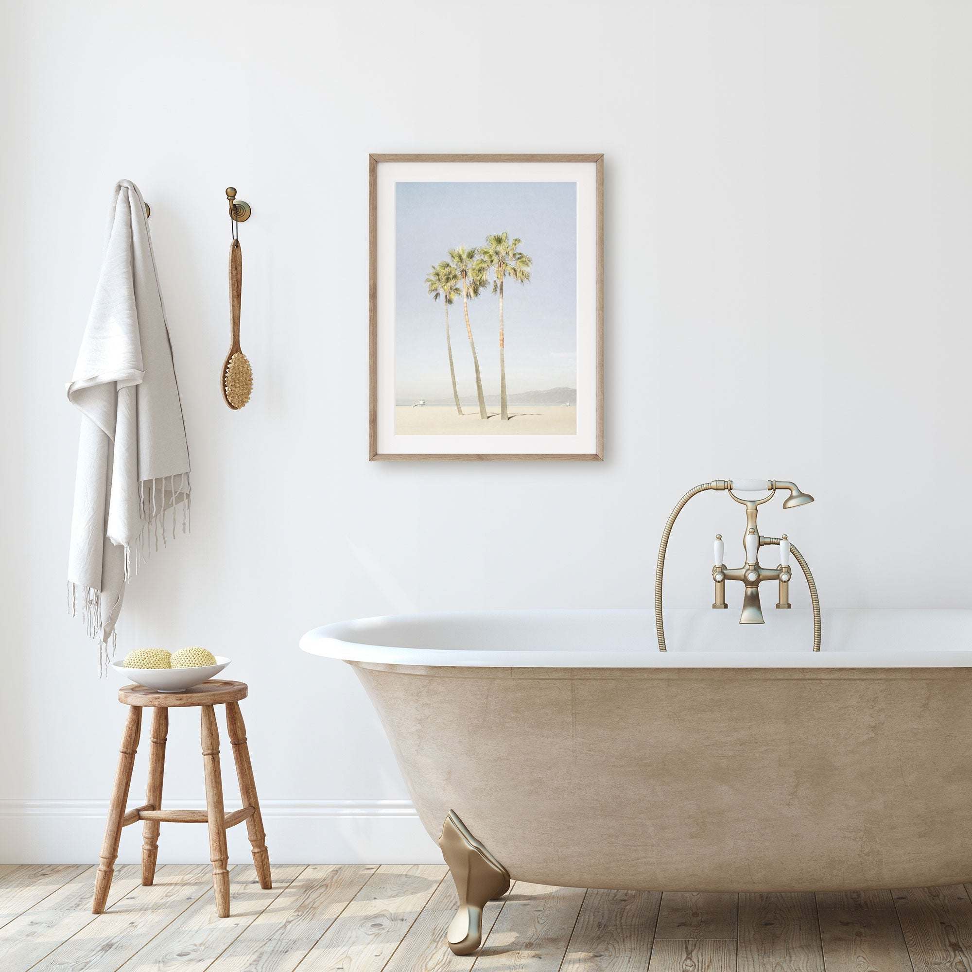 A minimalist bathroom featuring a clawfoot bathtub with golden fixtures, a wooden stool with bath items, and an unframed Offley Green California Venice Beach Print, 'Three Palms' on the wall next to hanging towels.