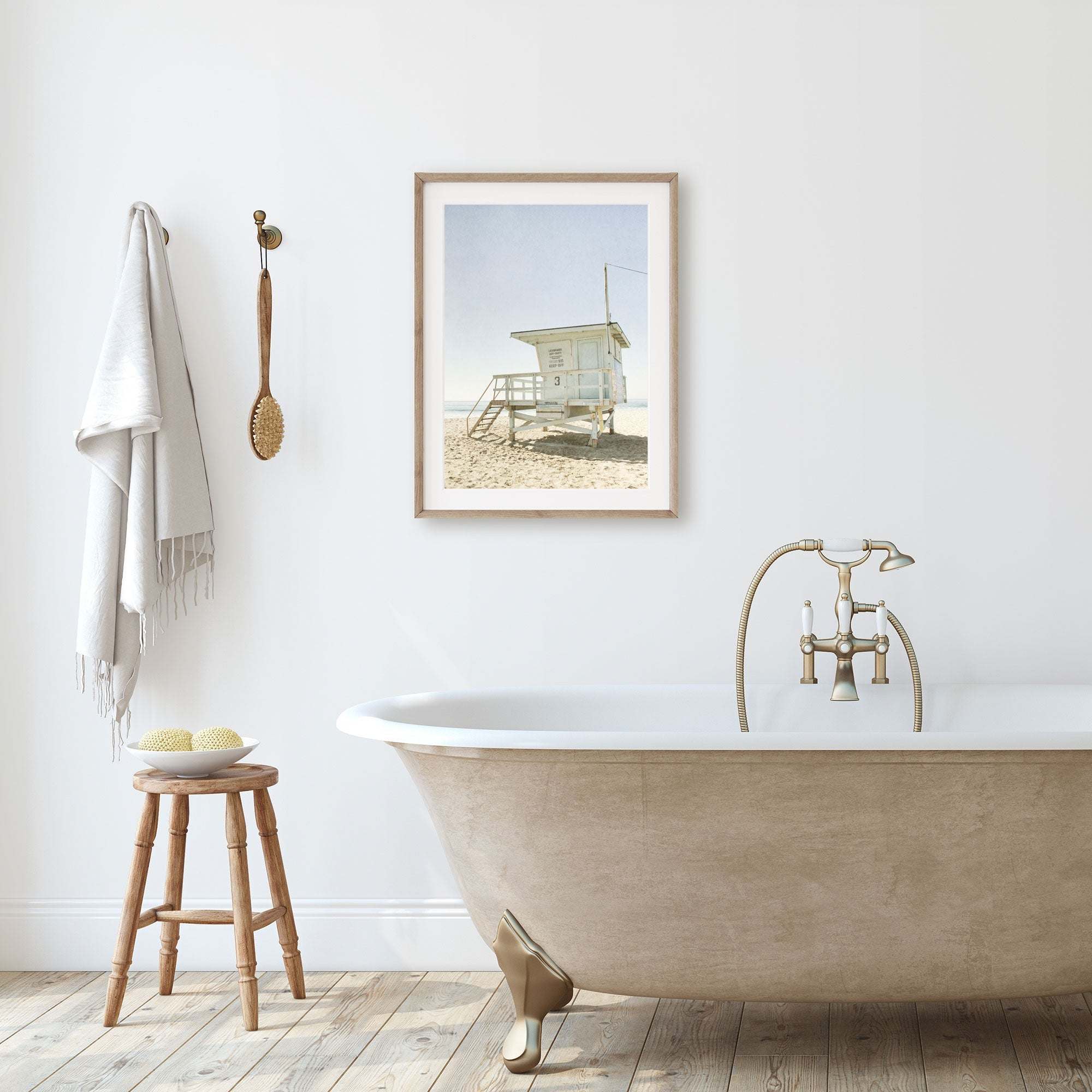 A minimalist bathroom with a classic clawfoot tub near a wooden stool with sponges, next to a wall with an unframed California Malibu Beach Print, 'Surfrider Lifeguard' photograph, and towels hanging on hooks by Offley Green.