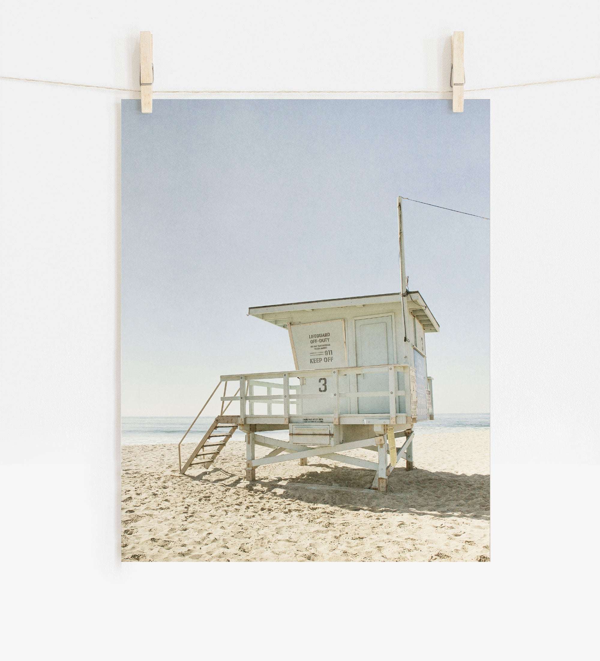 A photo of a California Malibu Beach print, 'Surfrider Lifeguard' printed on archival photographic paper, hanging against a sky-blue background, set on a sandy beach under bright sunlight. (Offley Green)