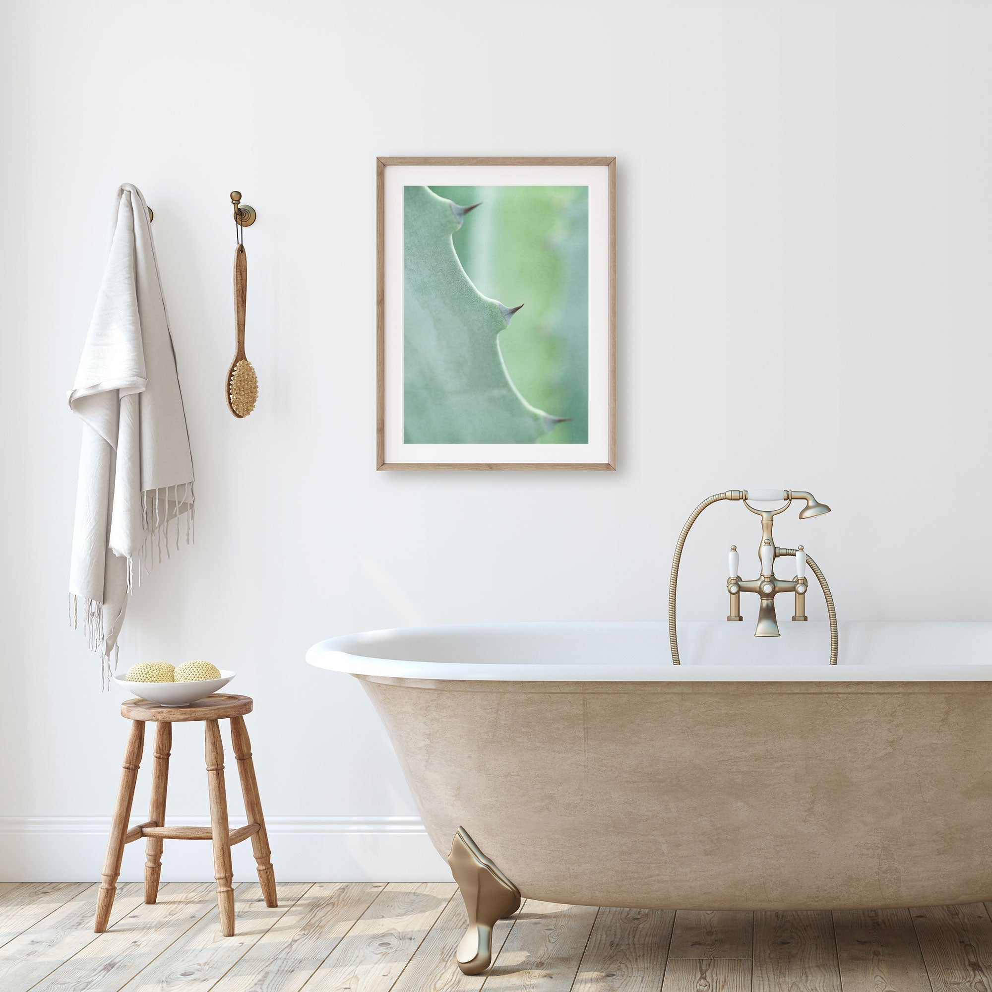 A minimalistic bathroom featuring a freestanding bathtub with golden claw feet, a wooden stool with bath items, a towel hanging on the wall, and Offley Green&#39;s &#39;Aloe Vera Spikes II&#39; unframed print above the tub.