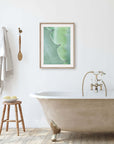 A minimalistic bathroom featuring a freestanding bathtub with golden claw feet, a wooden stool with bath items, a towel hanging on the wall, and Offley Green's 'Aloe Vera Spikes II' unframed print above the tub.