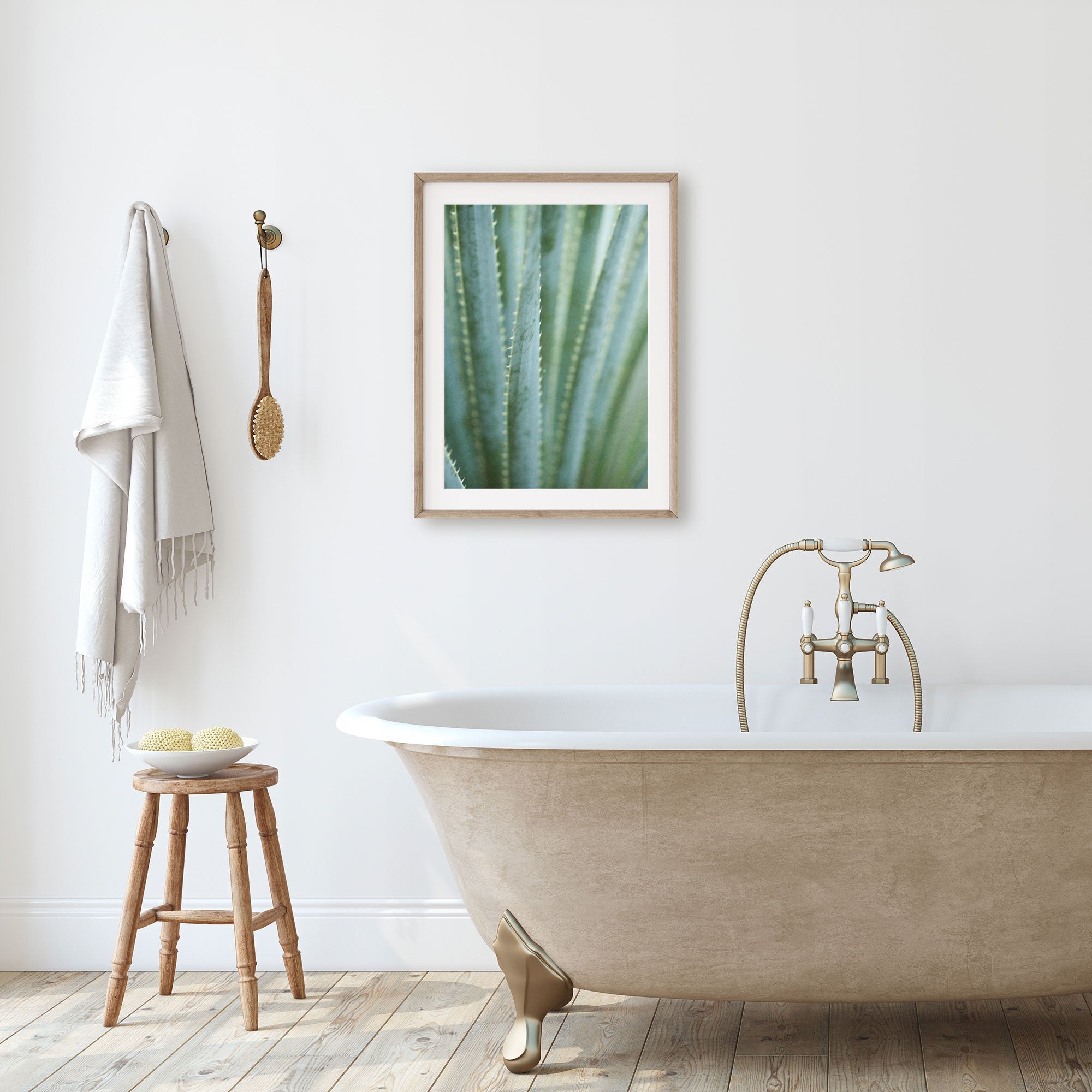 A minimalist bathroom with a freestanding bathtub, a stool holding bath accessories, and an unframed 'Strands and Spikes II' desert plants photograph by Offley Green on the wall. A towel hangs beside a wooden brush.