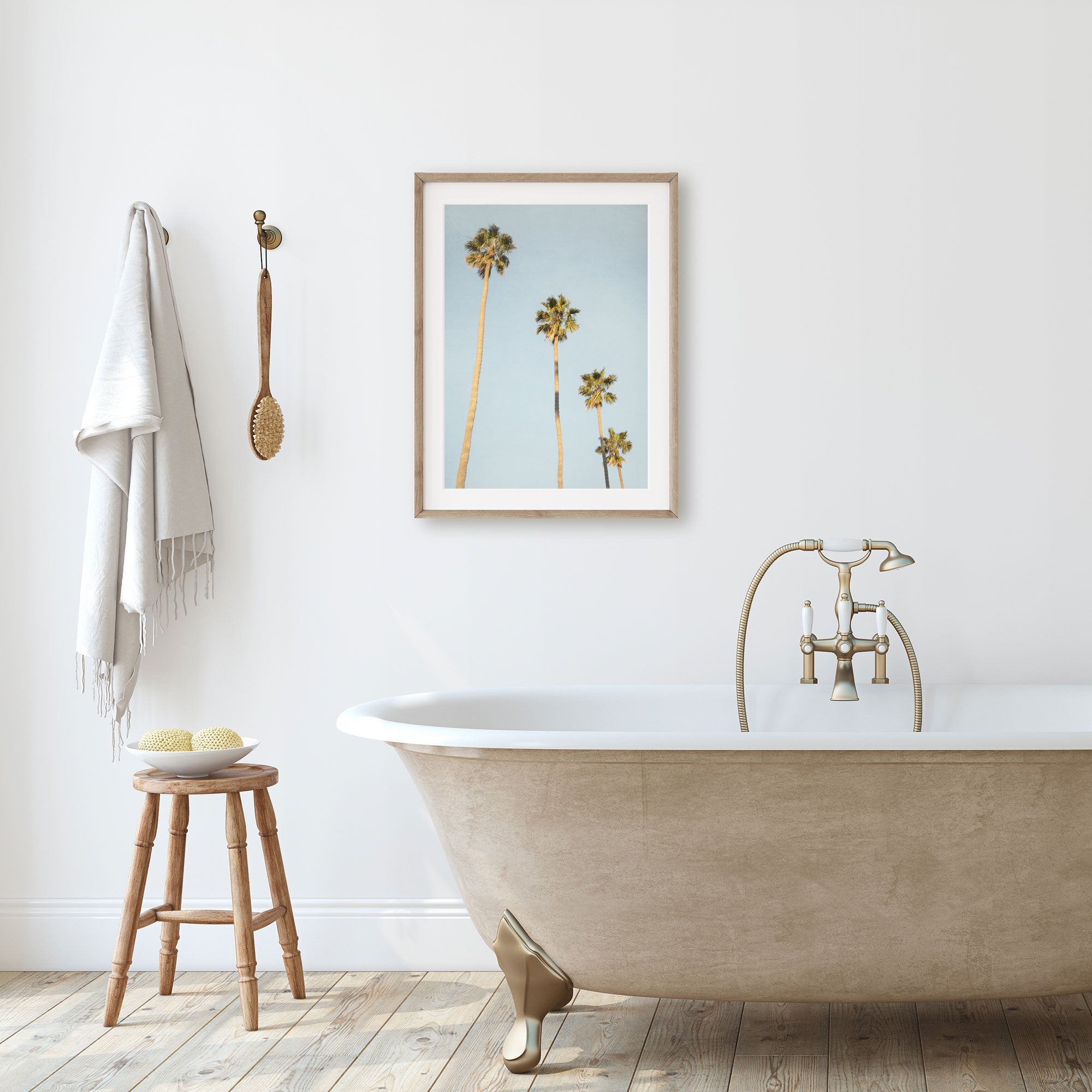 A minimalist bathroom featuring a standalone bathtub with gold clawfoot detail, a wooden stool, hanging towels, and a framed Offley Green Los Angeles Palm Tree Photographic Print 'Palm Stairs to Heaven' on the wall.