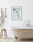 A minimalist bathroom featuring a standalone bathtub with gold clawfoot detail, a wooden stool, hanging towels, and a framed Offley Green Los Angeles Palm Tree Photographic Print 'Palm Stairs to Heaven' on the wall.
