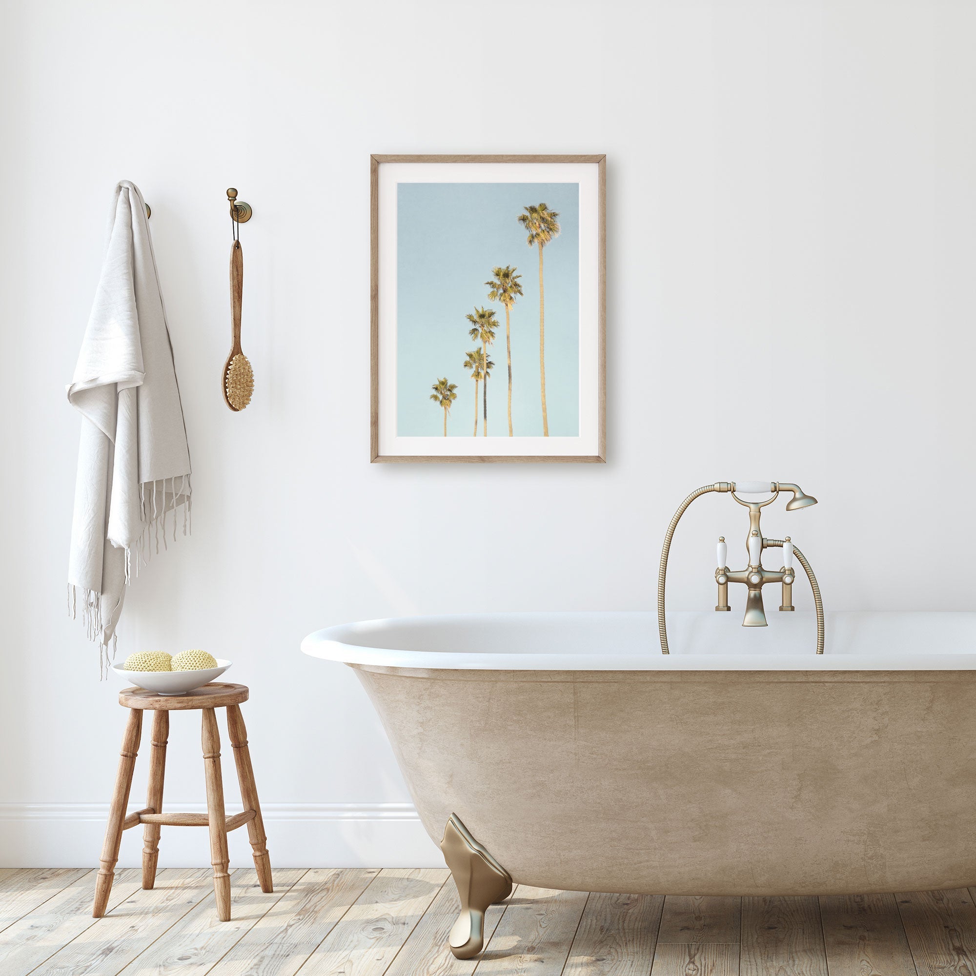 A minimalist bathroom featuring a clawfoot tub with a gold faucet, a wooden stool with toiletries, and a framed Offley Green "Los Angeles Palm Tree Photographic Print 'Palm Tree Steps'" on the wall, complemented by hanging towels.