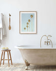 A minimalist bathroom featuring a clawfoot tub with a gold faucet, a wooden stool with toiletries, and a framed Offley Green "Los Angeles Palm Tree Photographic Print 'Palm Tree Steps'" on the wall, complemented by hanging towels.