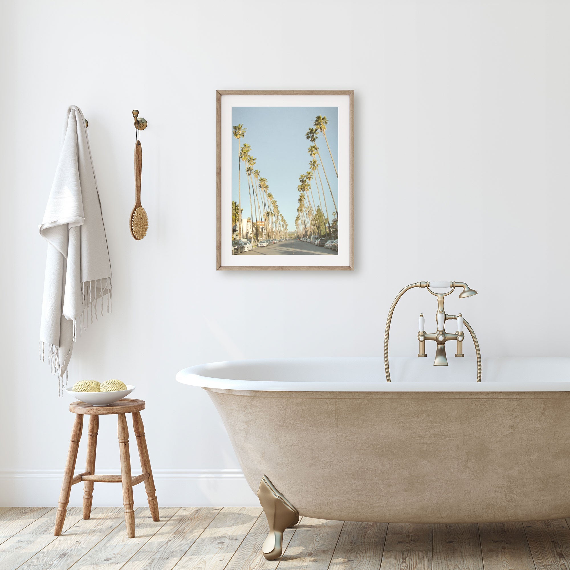 A minimalist bathroom featuring a clawfoot bathtub, golden faucets, a wooden stool with towels, and a framed painting of Los Angeles Palm Tree Lined Street &#39;Sunset Boulevard Dreams&#39; by Offley Green on the wall.