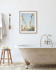 A minimalist bathroom featuring a clawfoot bathtub, golden faucets, a wooden stool with towels, and a framed painting of Los Angeles Palm Tree Lined Street 'Sunset Boulevard Dreams' by Offley Green on the wall.