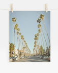 A photograph of a street lined with tall palm trees on Sunset Boulevard under a clear blue sky, pinned up by two clothespins on a wire, against a white background featuring the Los Angeles Palm Tree Lined Street 'Sunset Boulevard Dreams' by Offley Green.