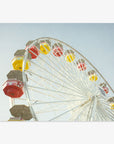 A colorful Santa Monica Ferris Wheel Print with red, yellow, and brown cabins against a clear blue sky at Santa Monica Pier by Offley Green.