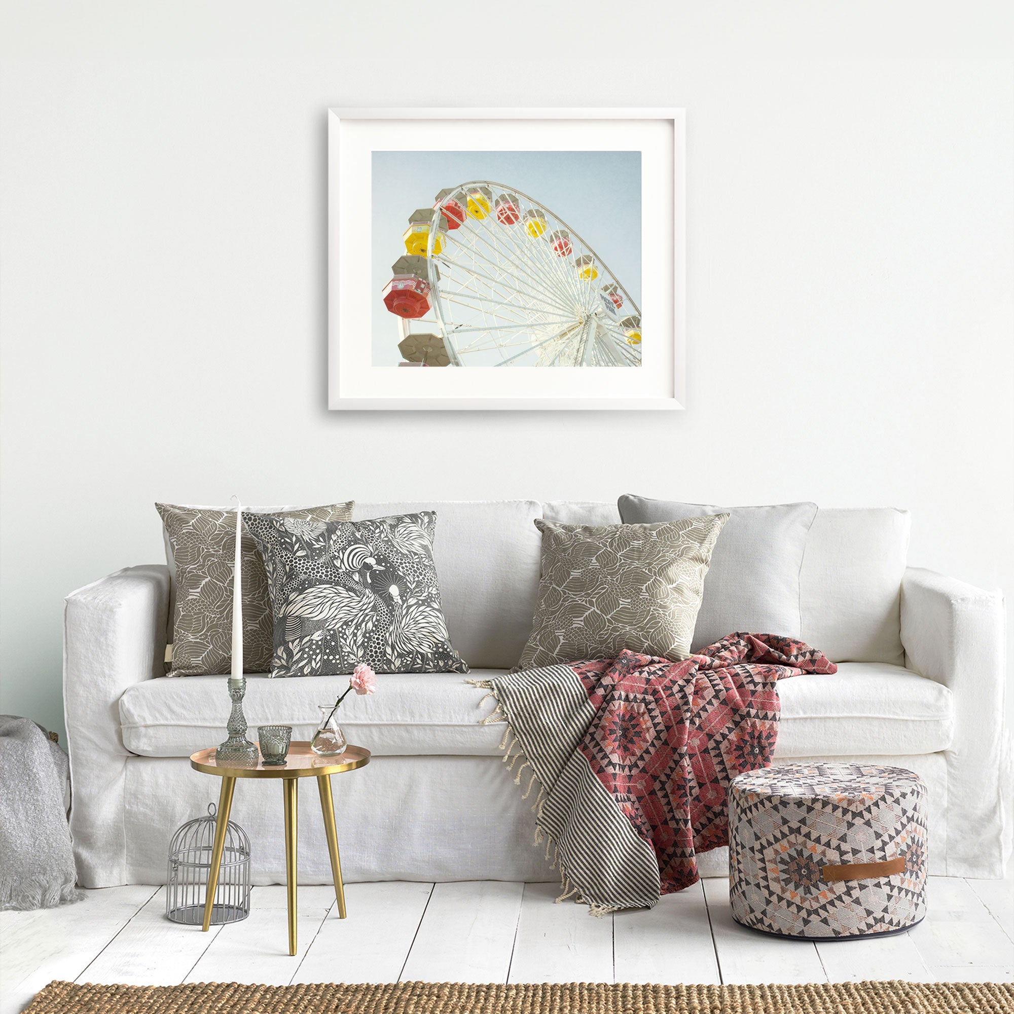 A cozy living room corner with a white sofa adorned with patterned cushions, a red throw blanket, and a small round gold table with a pink rose in a vase. A framed picture of the Offley Green Santa Monica Ferris Wheel Print, 'Ferris Above'