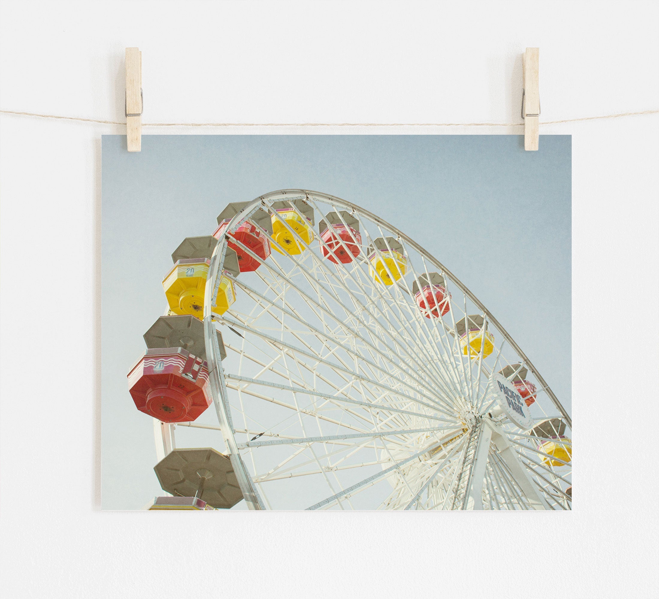 A photograph of a colorful Santa Monica Ferris Wheel Print, 'Ferris Above' by Offley Green is clipped to a string by wooden clothespins against a white wall. The sky is clear and blue in the background.