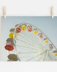 A photograph of a colorful Santa Monica Ferris Wheel Print, 'Ferris Above' by Offley Green is clipped to a string by wooden clothespins against a white wall. The sky is clear and blue in the background.