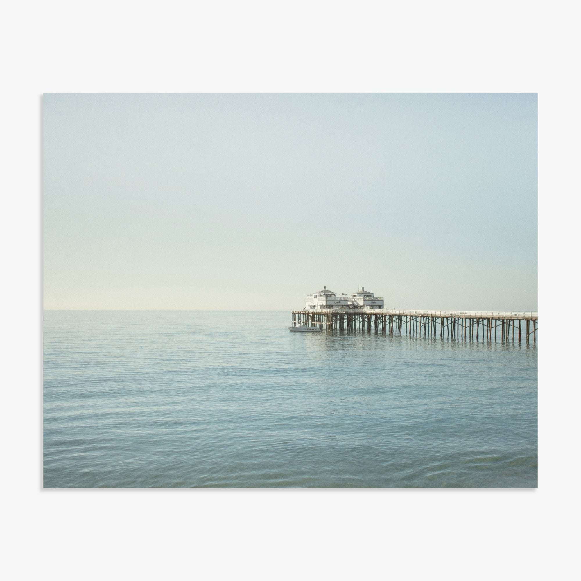 A tranquil seascape featuring Offley Green&#39;s &#39;Coastal Print of Malibu Pier in California &quot;All Calm in Malibu&quot; extending into calm waters with a large structure at the end under a clear sky.