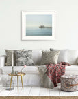 A cozy living room with a white sofa filled with decorative pillows, a red throw blanket, and a framed picture of Offley Green's Coastal Print of Malibu Pier in California 'All Calm in Malibu' above it. A small table with a book and a flower.
